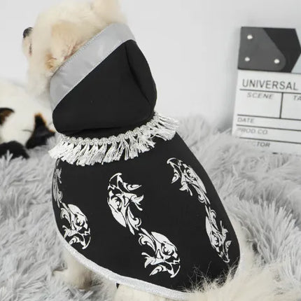 Small white dog wearing a black Halloween cape with silver patterns and fringe.