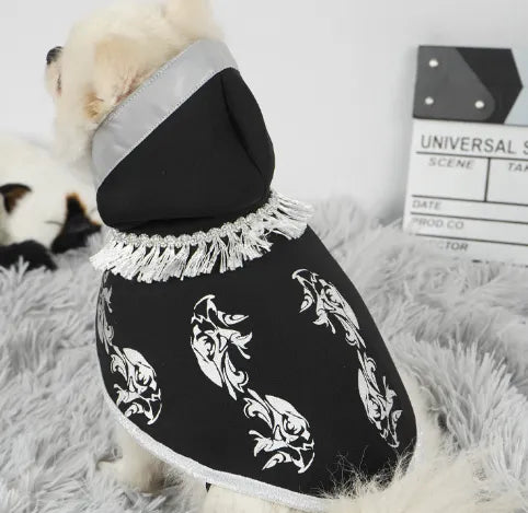 Small white dog wearing a black Halloween cape with silver patterns and fringe.